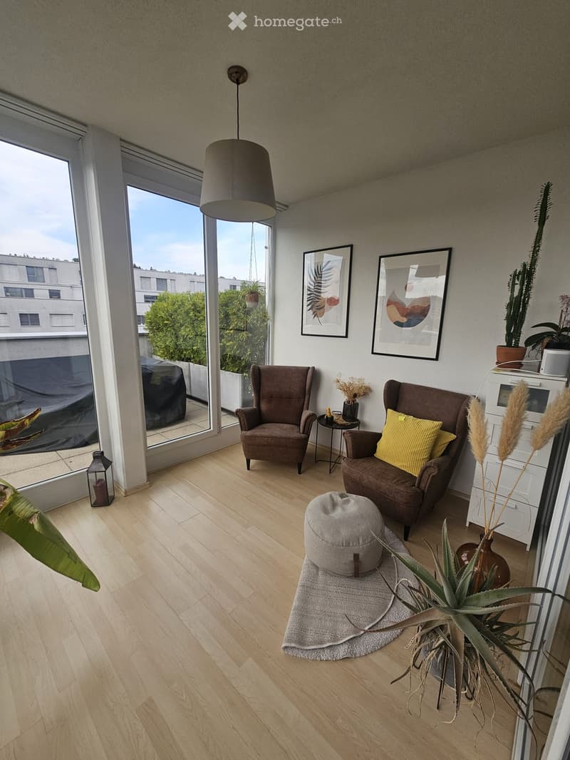 Modern living room with light wooden flooring, large windows, two armchairs, an ottoman, a small side table, and decorative plants.