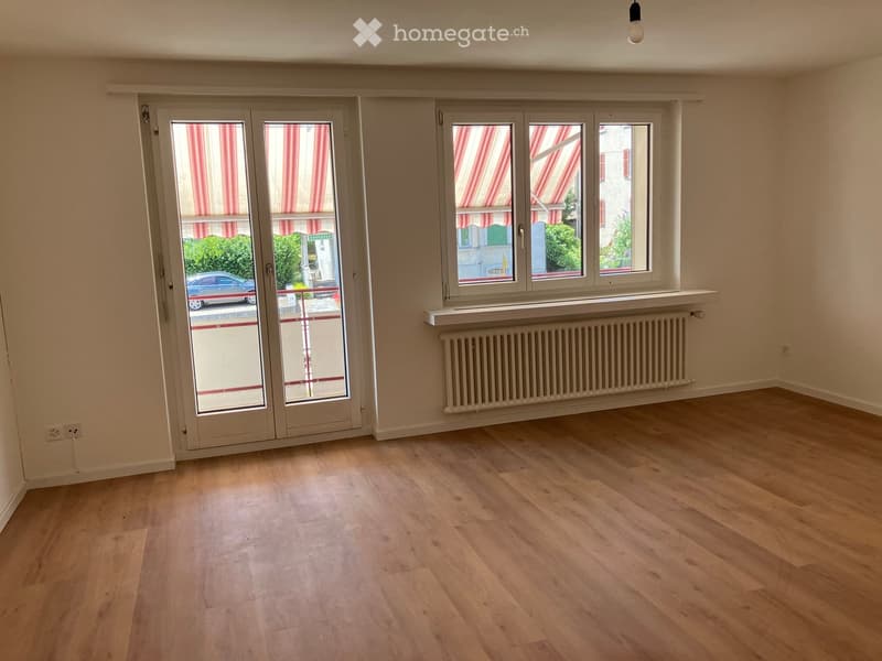 Empty room with light wooden flooring, large windows with striped blinds, and a radiator under one window.
