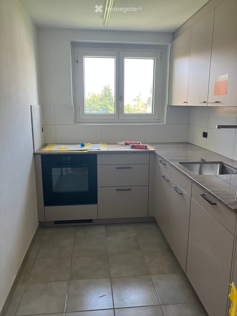 Modern kitchen with grey cabinets, tiled floor, and a window above the sink. The oven is installed under the countertop, and the sink is on the right side.