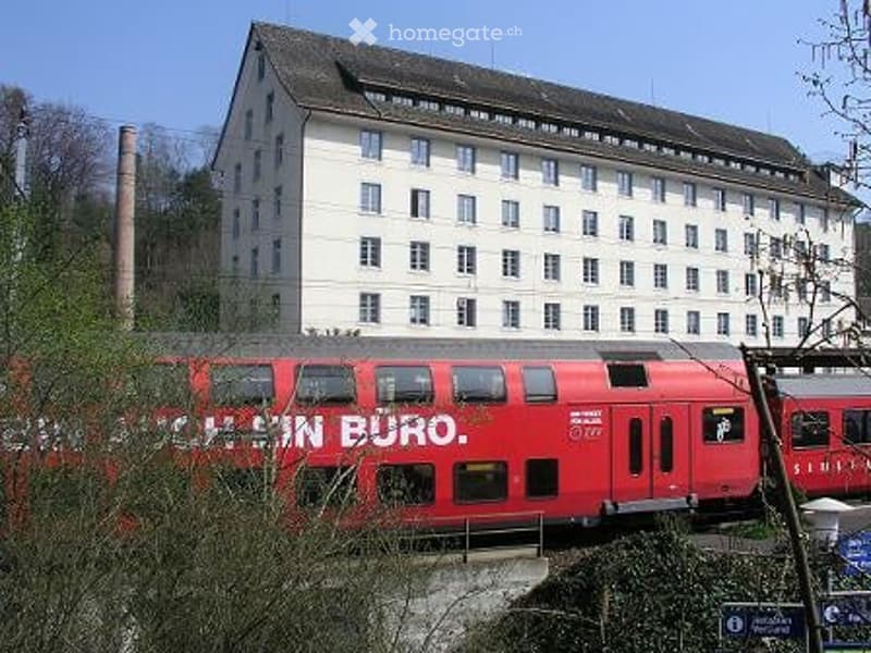 A large, multi-story building with white walls and numerous windows, situated near a railway track with a red train passing by.