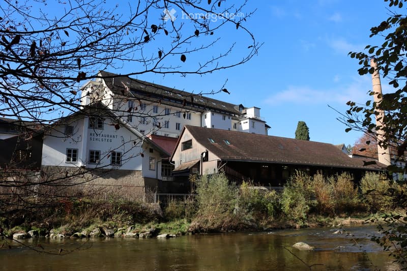 A multi-story building with a restaurant sign, situated near a river with a stone embankment and surrounding greenery.