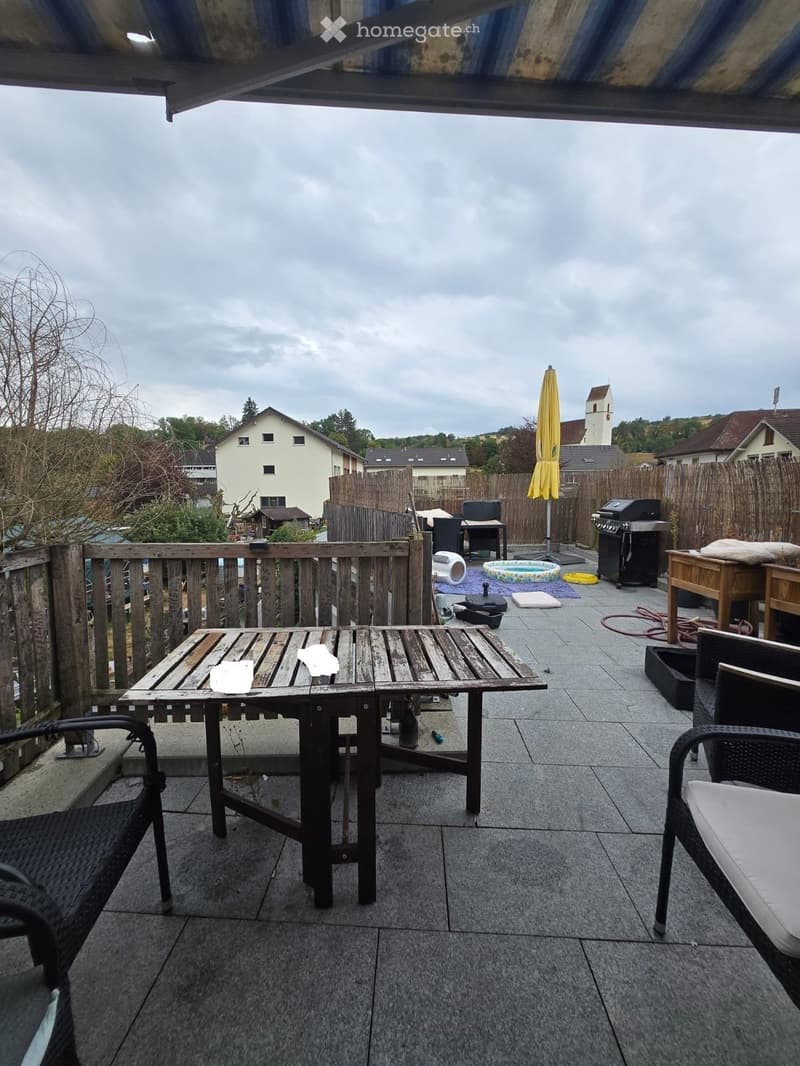Wooden table and chairs on a tiled balcony, with a closed umbrella and grill in the background, overlooking a residential area.