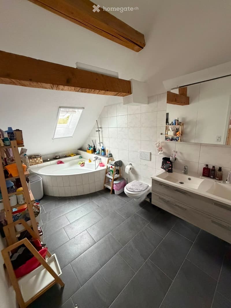 Modern bathroom with a large bathtub, toilet, sink, and mirror. Features wooden beams on the ceiling and a window with blinds. Shelves hold various toiletries and towels.