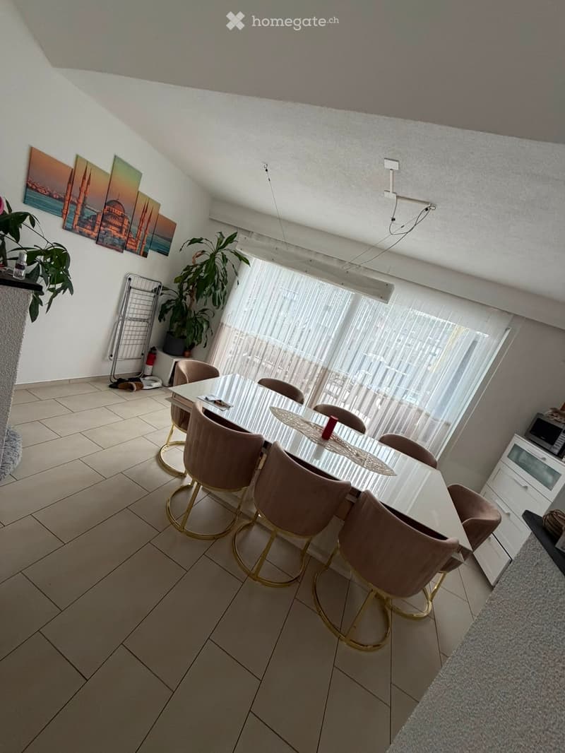 Modern dining area with a round glass table, six upholstered chairs, large windows with sheer curtains, and a potted plant near the window. The room features tiled flooring and white walls adorned with artwork.