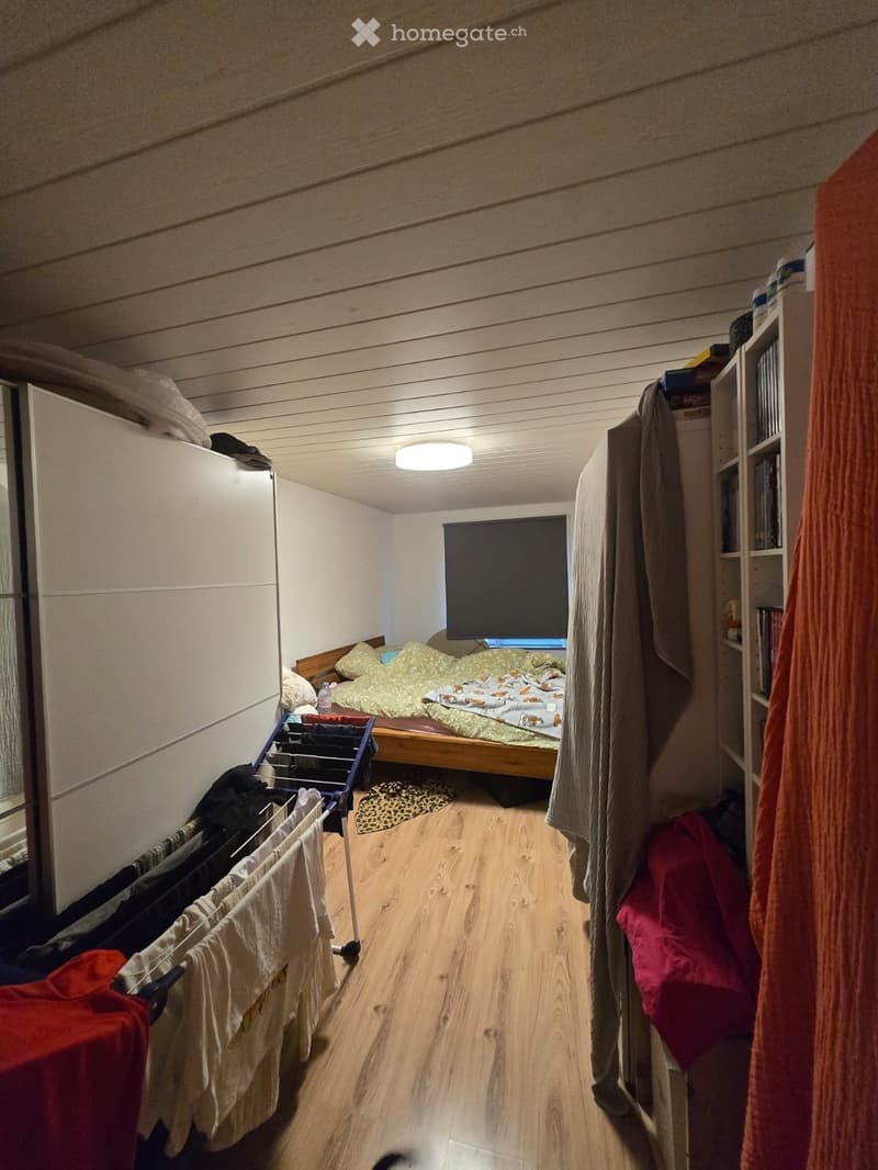 Cozy bedroom with wooden flooring, a single bed with colorful bedding, a bookshelf, a clothing rack, and a mirror. The room features a small window and a ceiling light.