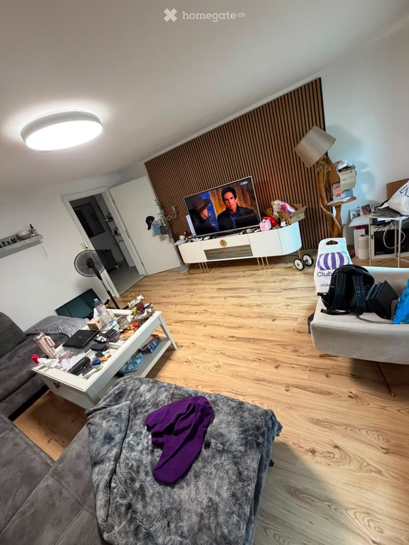 Living room with wooden flooring, a grey sofa, a coffee table, a television, and a fan. There is also a purple blanket on the sofa and a backpack on the floor.