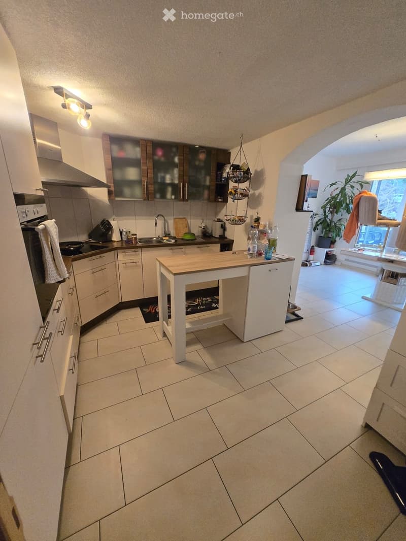 Modern kitchen with white cabinets, a central island, and stainless steel appliances. The space features a tiled floor and is well-lit with recessed lighting. A small living area is visible through an open doorway.