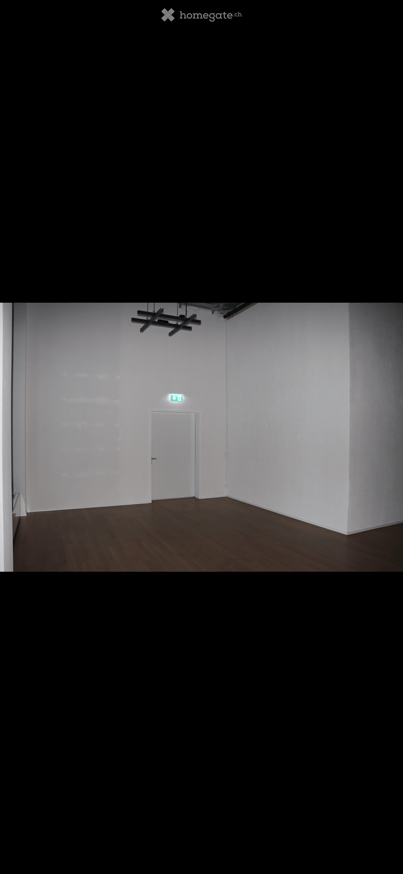 Empty room with white walls, wooden floor, and a single white door with an emergency exit sign above it.