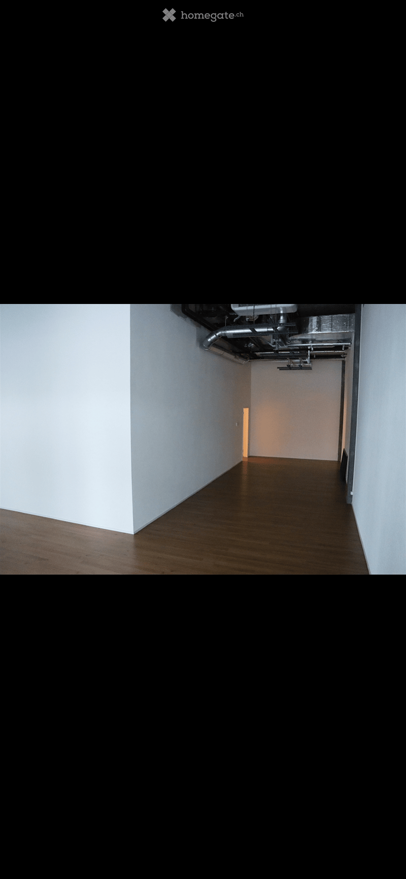 Empty hallway with wooden flooring and white walls, leading to a room with a visible door and a small light fixture.