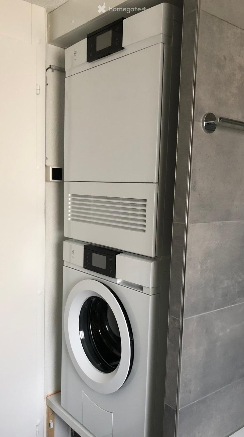 A corner of a room with two white appliances stacked on top of each other, a modern dryer on top and a washing machine below.