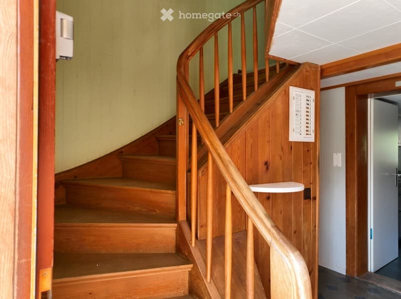 Wooden staircase with handrail, light-colored walls, and a small white shelf on the wall.