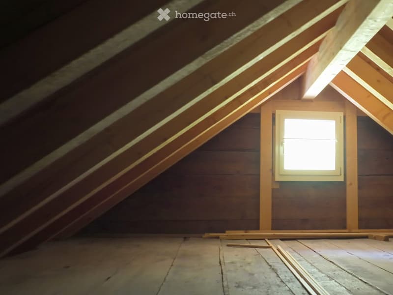 Empty attic space with wooden walls, exposed beams, and a single window letting in natural light. The floor is unfinished and shows some wear.