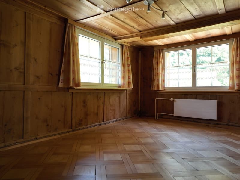 Empty room with wooden walls, wooden floor, large windows with curtains, and a radiator.