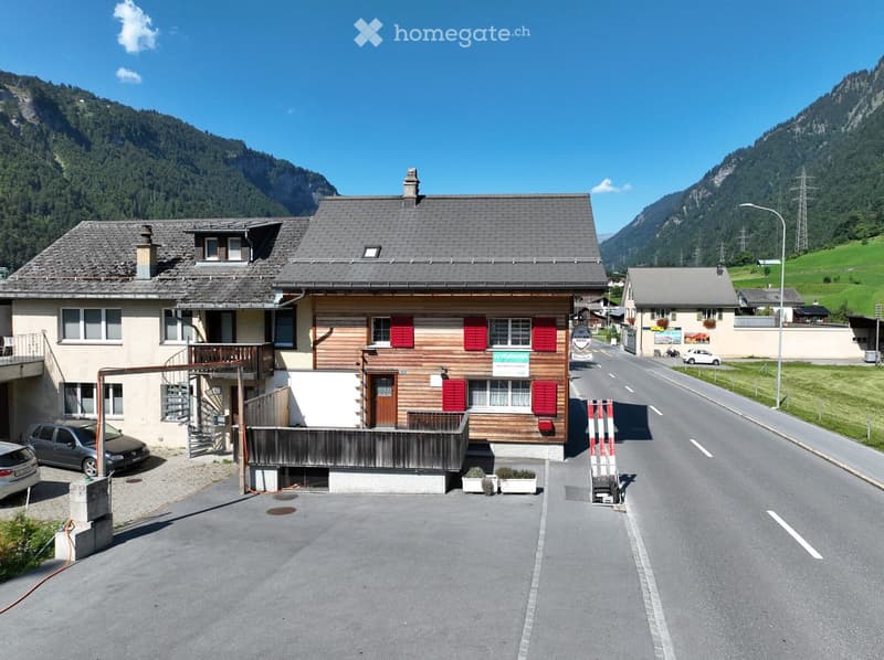 Two-story wooden house with red shutters, a balcony, and a driveway with parked cars. Surrounded by green hills and mountains.