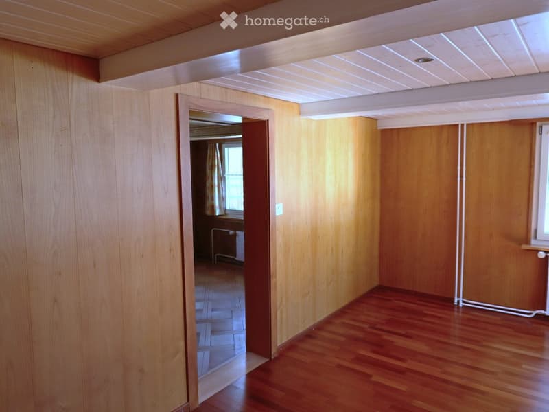 Empty room with wooden walls and polished wooden floor, featuring a doorway leading to another room with a window and radiator.
