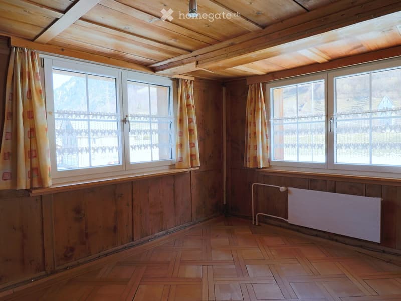 Empty room with wooden walls, wooden ceiling, wooden floor, large windows with curtains, radiator, and visible beams on the ceiling.