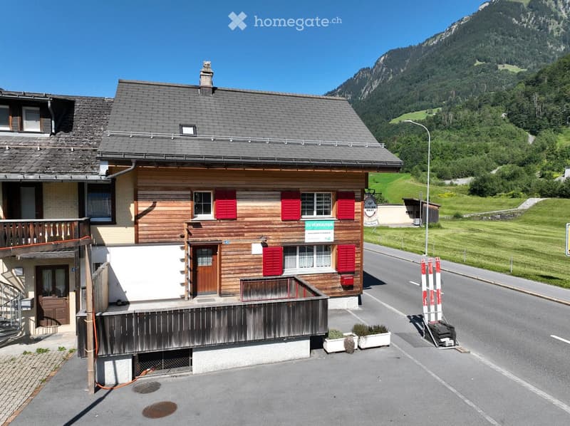 Two-story wooden house with red shutters, a balcony, and a view of mountains in the background.