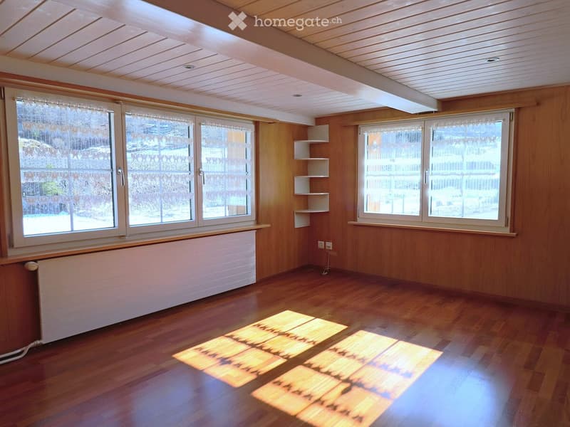 Empty room with wooden walls, wooden floor, large windows with lace curtains, radiator, and built-in shelves.