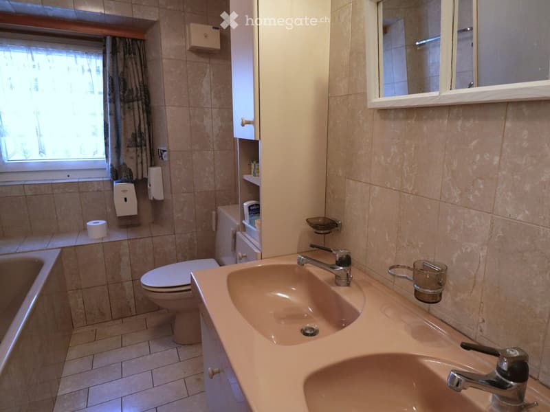 Bathroom with beige tiles, double sink, toilet, mirror, and cabinet. Includes a window with a curtain and a toilet paper holder.