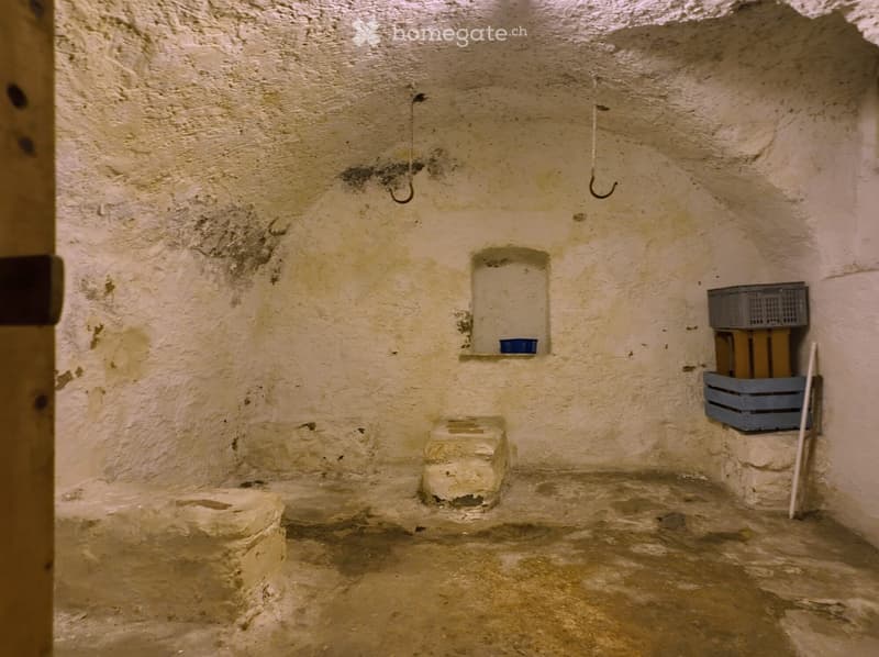Empty room with rough, textured stone walls and a dirt floor. There are two stone stools, a small shelf with a blue container, and a stack of crates with a broom leaning against the wall.