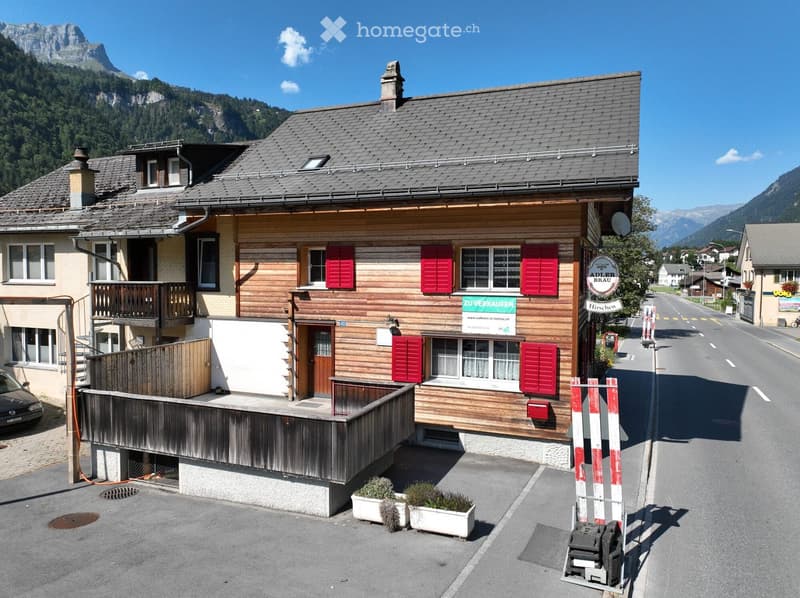 Two-story wooden house with red shutters, a balcony, and a sign reading "Zu Verkaufen." The house is situated on a street with mountains in the background.