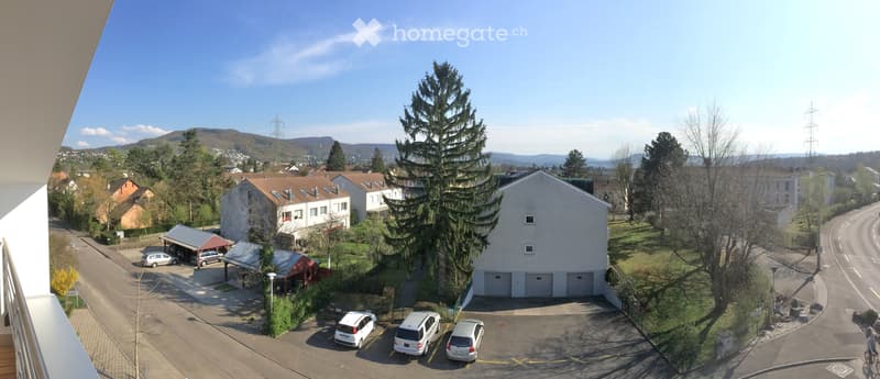 View from a balcony showing a residential area with houses, garages, parked cars, and a scenic mountain backdrop.
