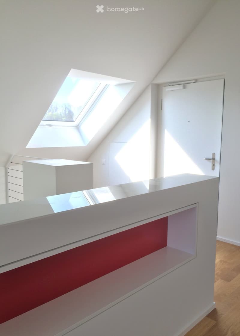 Bright, modern attic space with wooden flooring, white walls, and skylight windows allowing natural light. Features a white door and a sleek white counter with a red accent panel.