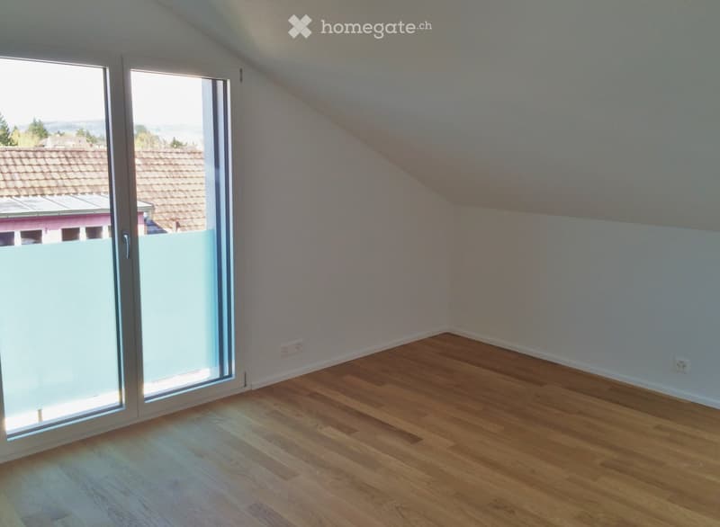 Empty attic room with wooden flooring, white walls, and large sliding glass doors leading to a balcony with a view of the surrounding area.