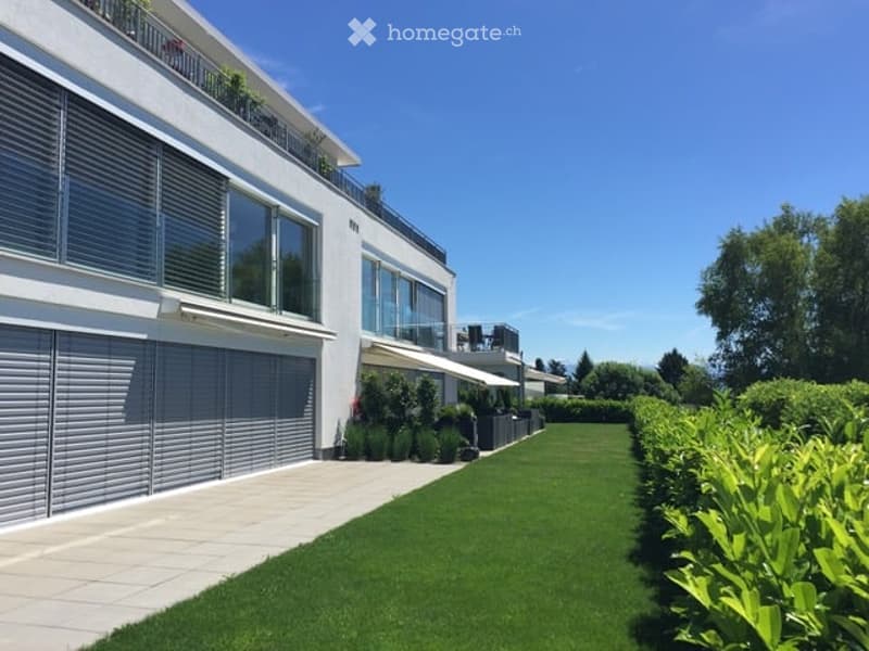 Modern white building with large windows, balconies, and a well-maintained lawn and hedge border.