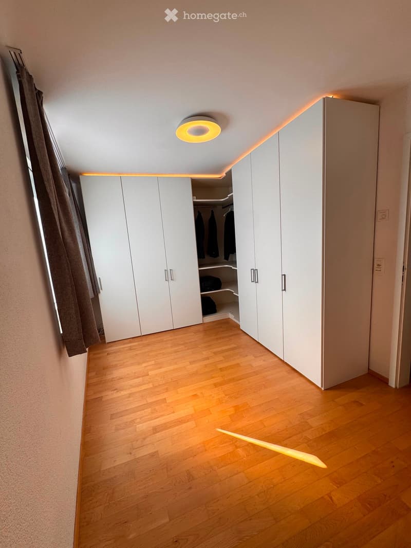 Empty room with wooden flooring, white built-in wardrobes, and a round ceiling light fixture. The room features a window with a curtain on the left side.