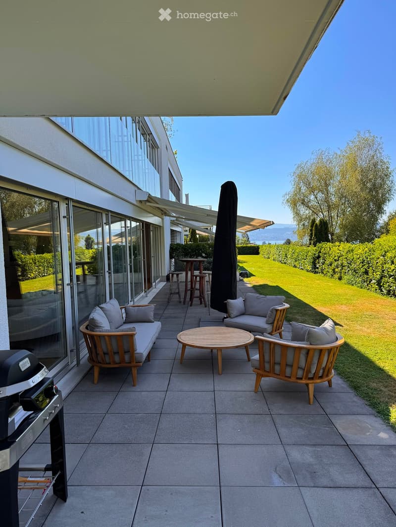 Balcony with a sofa, chairs, a round table, and an umbrella, overlooking a well-maintained lawn and garden.
