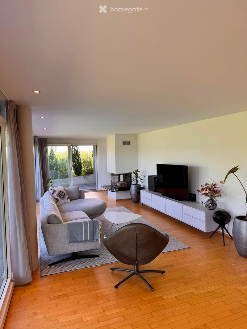 Modern living room with wooden flooring, large windows, grey sectional sofa, armchair, coffee table, TV, fireplace, and decorative plants.