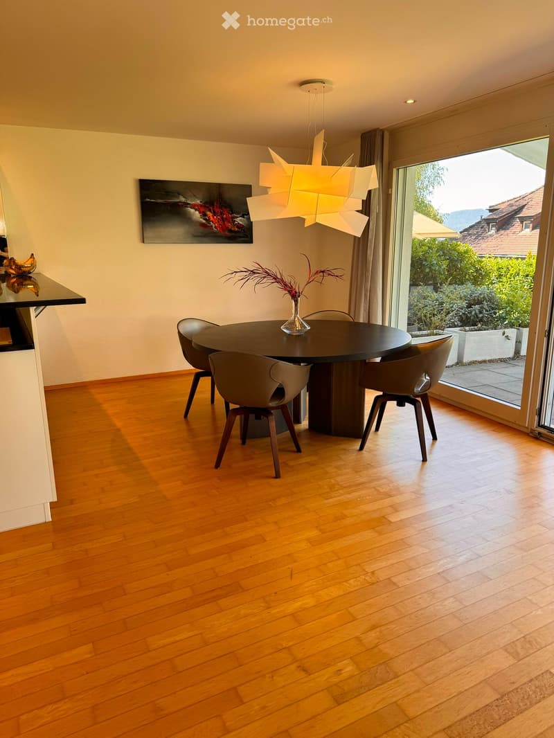 Dining room with wooden floor, round dining table, four chairs, vase with dried flowers, modern chandelier, and artwork on the wall. Large window offers a view of the garden.