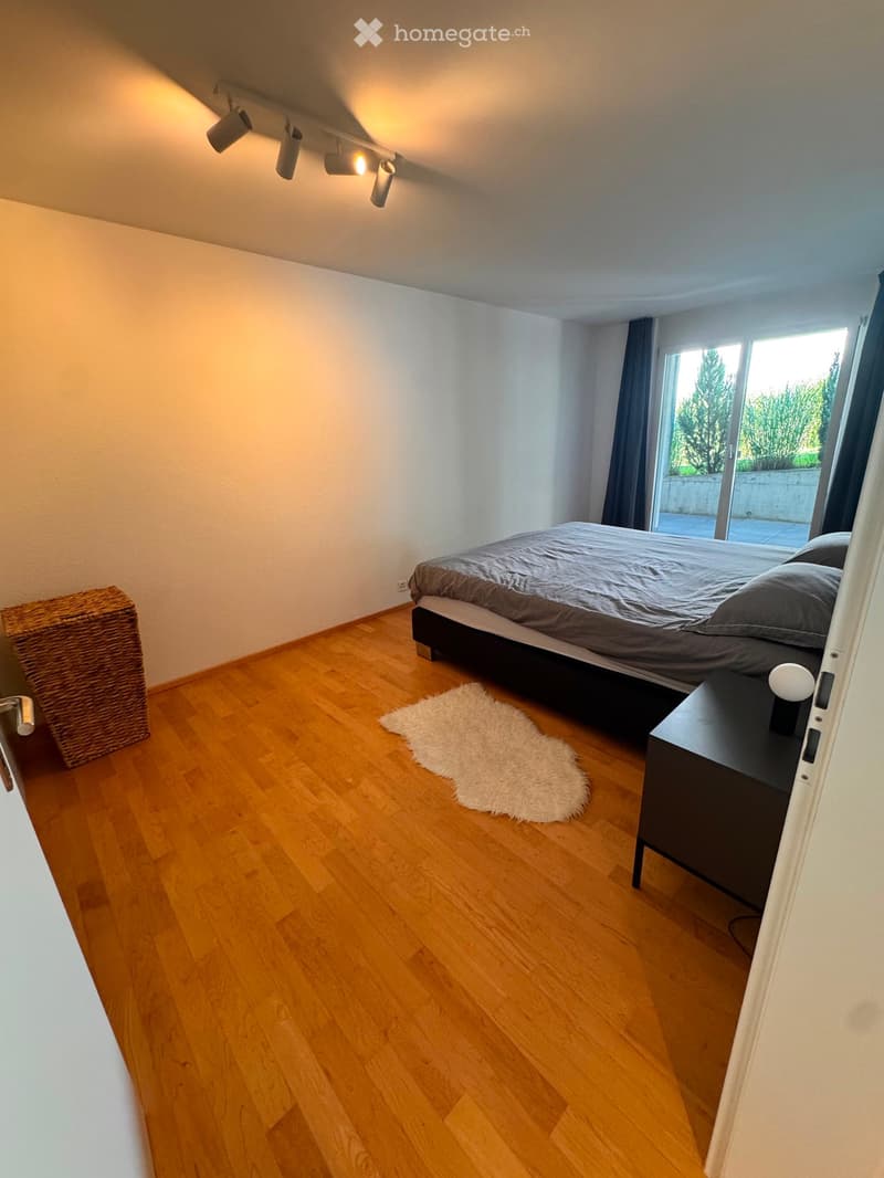 Modern bedroom with wooden flooring, a bed with grey bedding, a nightstand with a lamp, a white shag rug, and a wicker basket near the wall.
