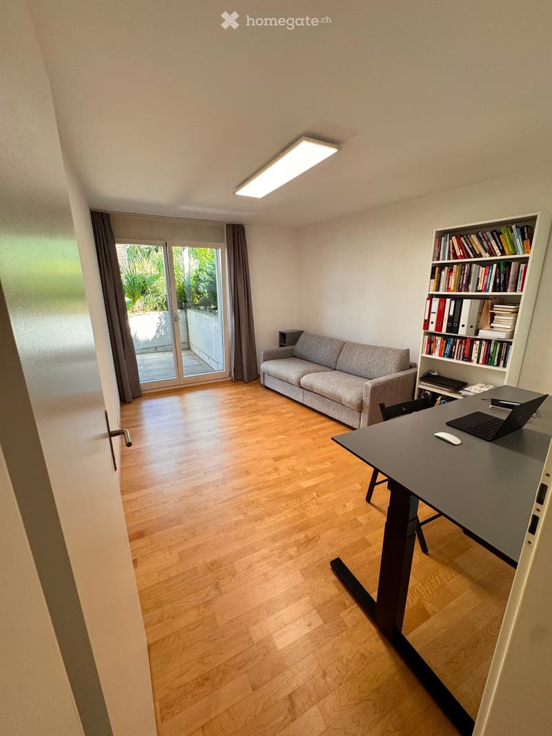 A modern, well-lit room with wooden flooring, a grey sofa, a desk with a laptop, a bookshelf filled with books, and a sliding glass door leading to a balcony.
