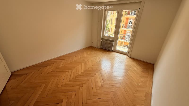 Empty room with wooden herringbone flooring, large window with sliding glass doors, radiator, and neutral-colored walls.