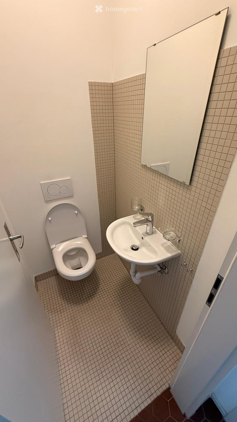Compact bathroom with a toilet, sink, and mirror. Beige tiled walls and floor, white fixtures.