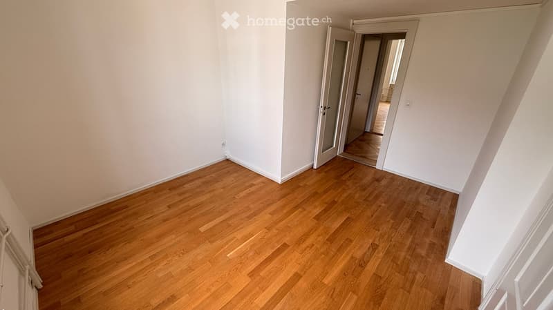 Empty room with light wooden flooring and white walls, featuring an open doorway leading to another room.