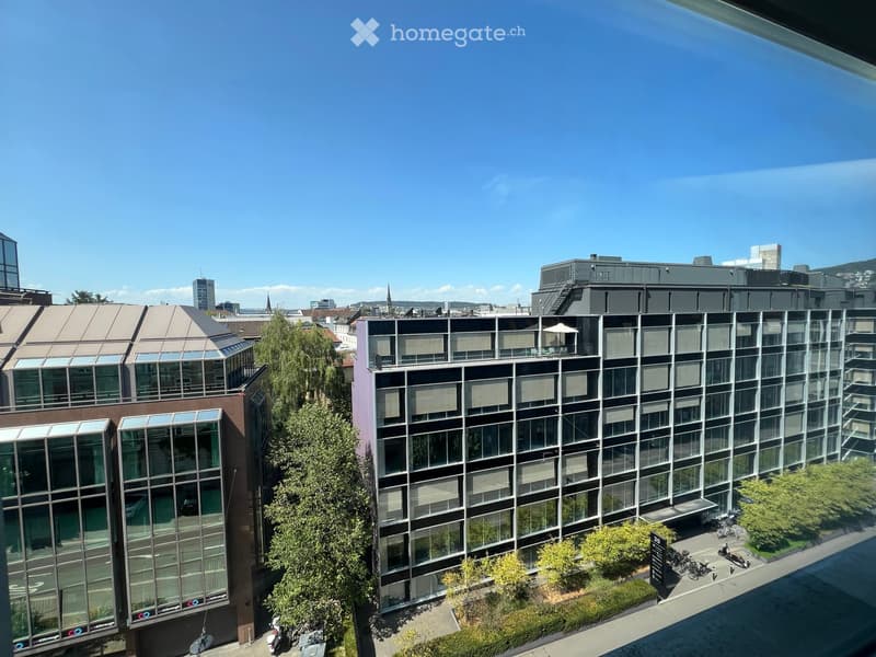 Modern multi-story building with large glass windows and balconies, surrounded by greenery and urban structures.