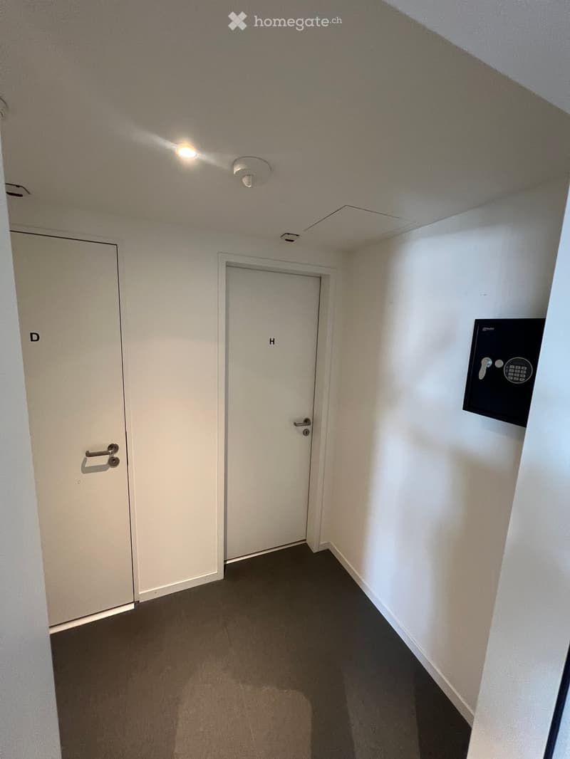 Empty hallway with two doors, one labeled 'D' and the other 'H', a security panel on the wall, and a ceiling light fixture.