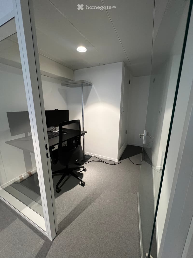 Modern office space with a grey carpeted floor, white walls, a black office chair, a desk with a computer and screen, and a glass partition.