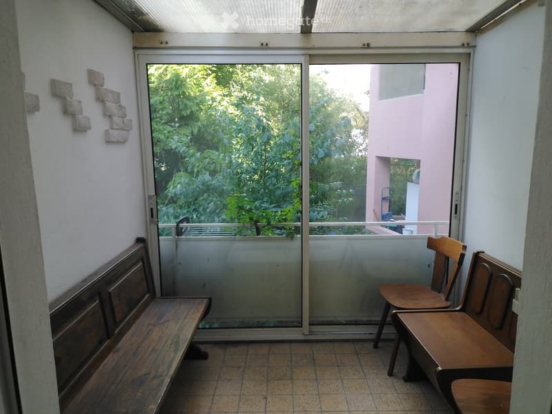 A small, enclosed balcony area with tiled flooring, a wooden bench on the left, and two wooden chairs on the right, with a large glass sliding door leading to a lush green outdoor view.