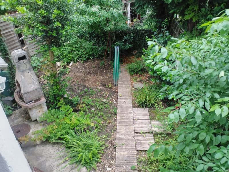 A lush garden with a wooden pathway, various plants, and a stone structure, likely a barbecue or fireplace, surrounded by greenery and trees.