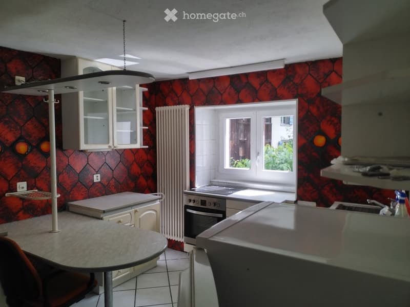 Kitchen with red patterned wallpaper, white cabinets, a round table, an oven, and a sink. Natural light comes through a window showing greenery outside.