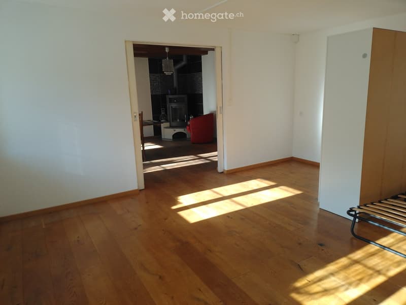 Bright, unfurnished room with wooden flooring and white walls, leading to a living area with a red sofa, fireplace, and a small table with chairs.