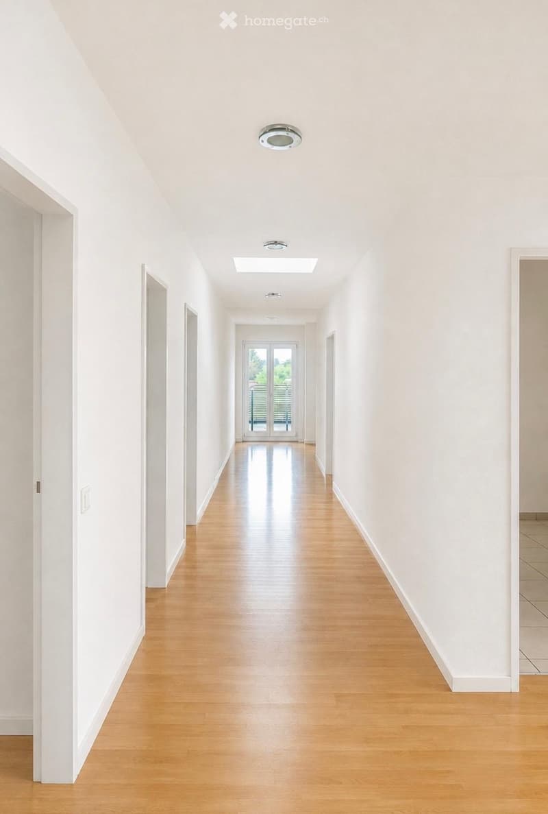 Bright, empty hallway with light wooden flooring, white walls, and recessed lighting. Features several doors and a skylight at the far end.
