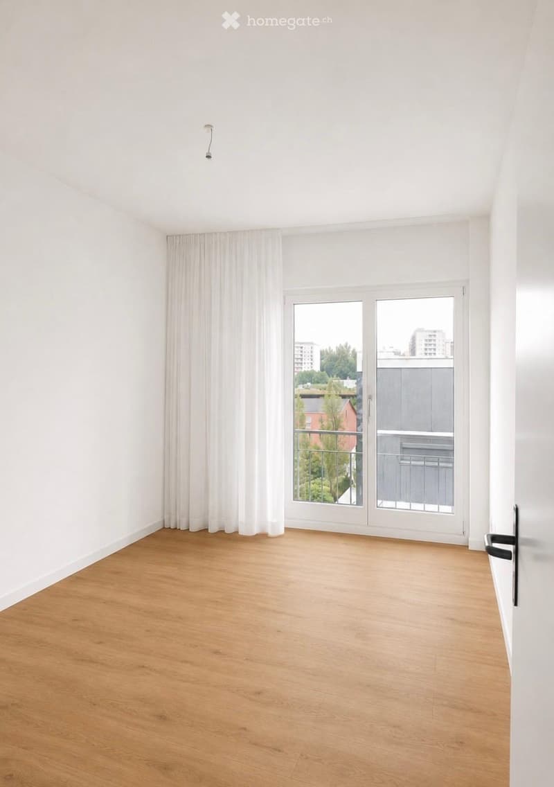 Empty room with light wooden flooring, white walls, large window with white curtains, and a sliding glass door leading to a balcony.