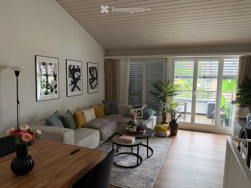 Modern living room with a grey sectional sofa, colorful cushions, a wooden coffee table, side tables, vases, and a floor lamp. Large windows and sliding doors lead to a balcony with outdoor furniture.