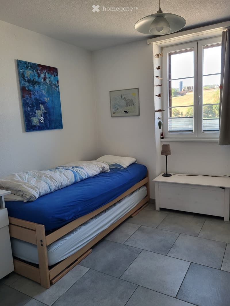 A small bedroom with a single bed, nightstand, lamp, and artwork on the walls. The room has tiled flooring and a window with blinds.