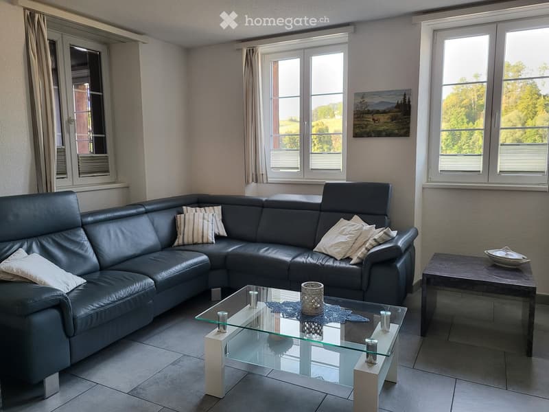 Modern living room with a dark leather sectional sofa, glass coffee table, side table, and decorative cushions. Large windows offer a scenic view.